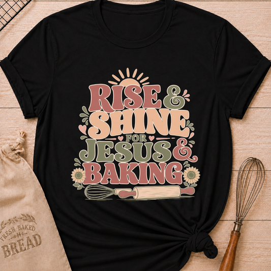 A black graphic t-shirt styled with a knotted hem, laid flat on a light wood background with a drawstring bread bag and a whisk. The shirt features a bold retro groovy graphic that reads "RISE & SHINE FOR JESUS & BAKING" in pink, cream, and sage lettering, with a rising sun at the top, heart accents, sunflowers, and illustrations of a whisk and rolling pin at the bottom.