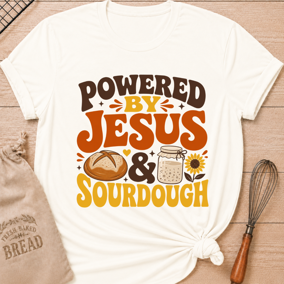 An off-white graphic t-shirt with a 'POWERED BY JESUS & SOURDOUGH' design, styled in a flat-lay on a wooden background with a burlap bag, bread, and a whisk. graphic t-shirt laid flat on a light wood background, styled with a bread bag, whisk, and baking paper. The t-shirt features a graphic that reads 'POWERED BY JESUS & SOURDOUGH' in brown and orange lettering, accompanied by illustrations of a sourdough bread loaf, a mason jar of dough, and a sunflower.