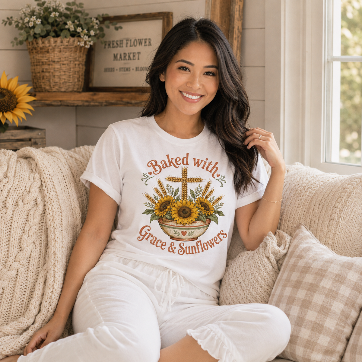 Smiling woman seated on a cozy couch wearing a white Baked with Grace & Sunflowers graphic tee featuring a cross, sunflowers, and wheat in a mixing bowl, with a sunflower and Fresh Flower Market sign in the background.