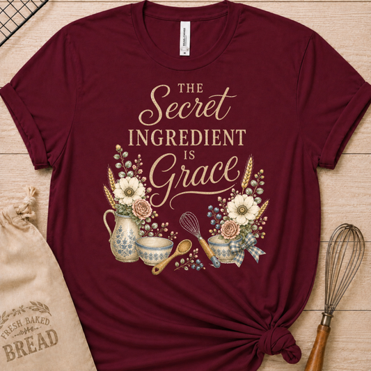 Maroon women's relaxed fit t-shirt reading The Secret Ingredient is Grace, framed by a floral pitcher, mixing bowls, a whisk, and a wooden spoon.