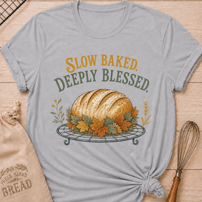 Athletic Heather women's relaxed fit tee with Slow Baked Deeply Blessed lettering over a golden artisan loaf resting on a vintage cooling rack, surrounded by richly colored fall leaves and wheat