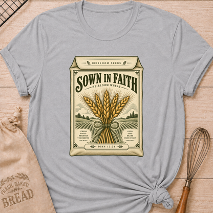 Athletic Heather women's relaxed fit tee with a vintage heirloom seed packet design, golden wheat, rolling fields and John 12:24 woven into the artwork beneath Sown in Faith lettering