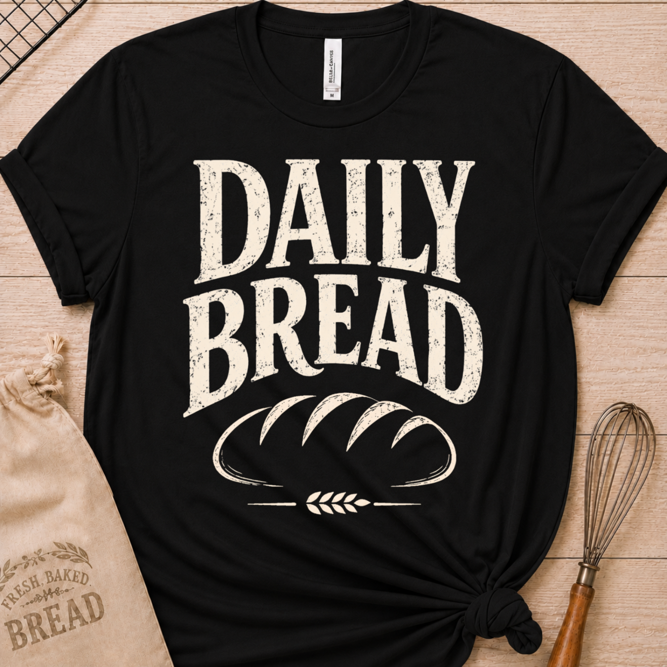 A black short-sleeve graphic T-shirt laid flat on a light wood surface, with large distressed white text reading “DAILY BREAD” and a bread loaf illustration on the front. A beige drawstring bag and a whisk are placed around it as styling props.