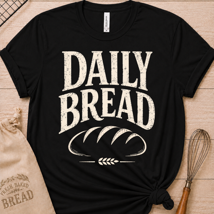 A black short-sleeve graphic T-shirt laid flat on a light wood surface, with large distressed white text reading “DAILY BREAD” and a bread loaf illustration on the front. A beige drawstring bag and a whisk are placed around it as styling props.