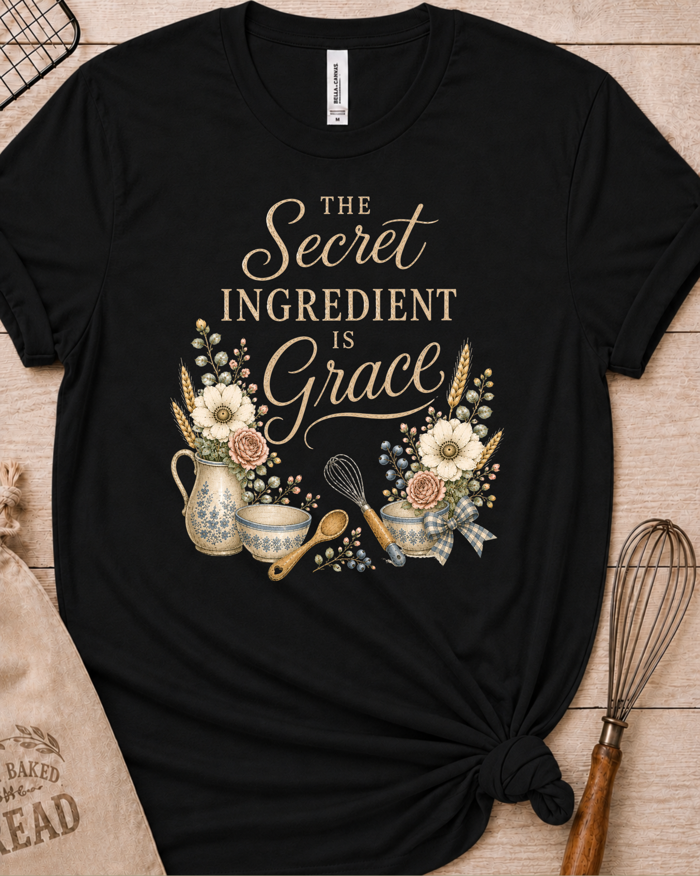 Black women's relaxed fit t-shirt reading The Secret Ingredient is Grace, framed by a floral pitcher, mixing bowls, a whisk, and a wooden spoon.