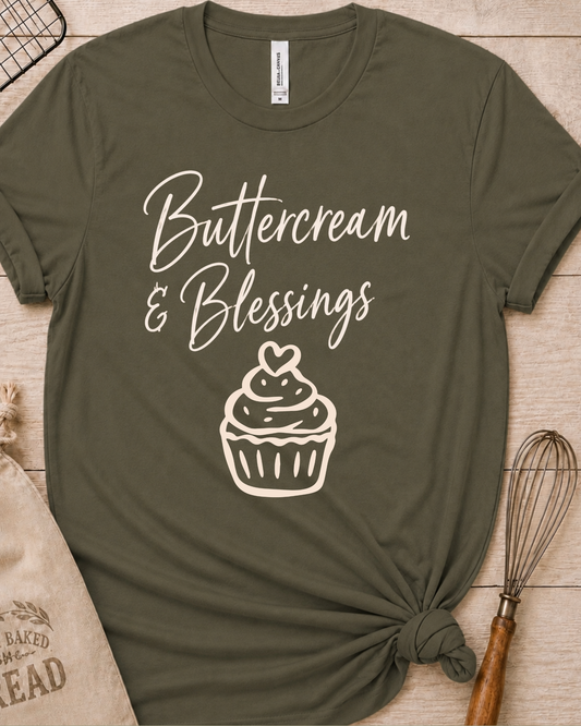 An olive green short-sleeve t-shirt with a knotted hem, featuring a white graphic print that reads 'Buttercream & Blessings' above a cupcake illustration. The shirt is styled flat on a light wood background with baking props, including a burlap sack and a whisk.