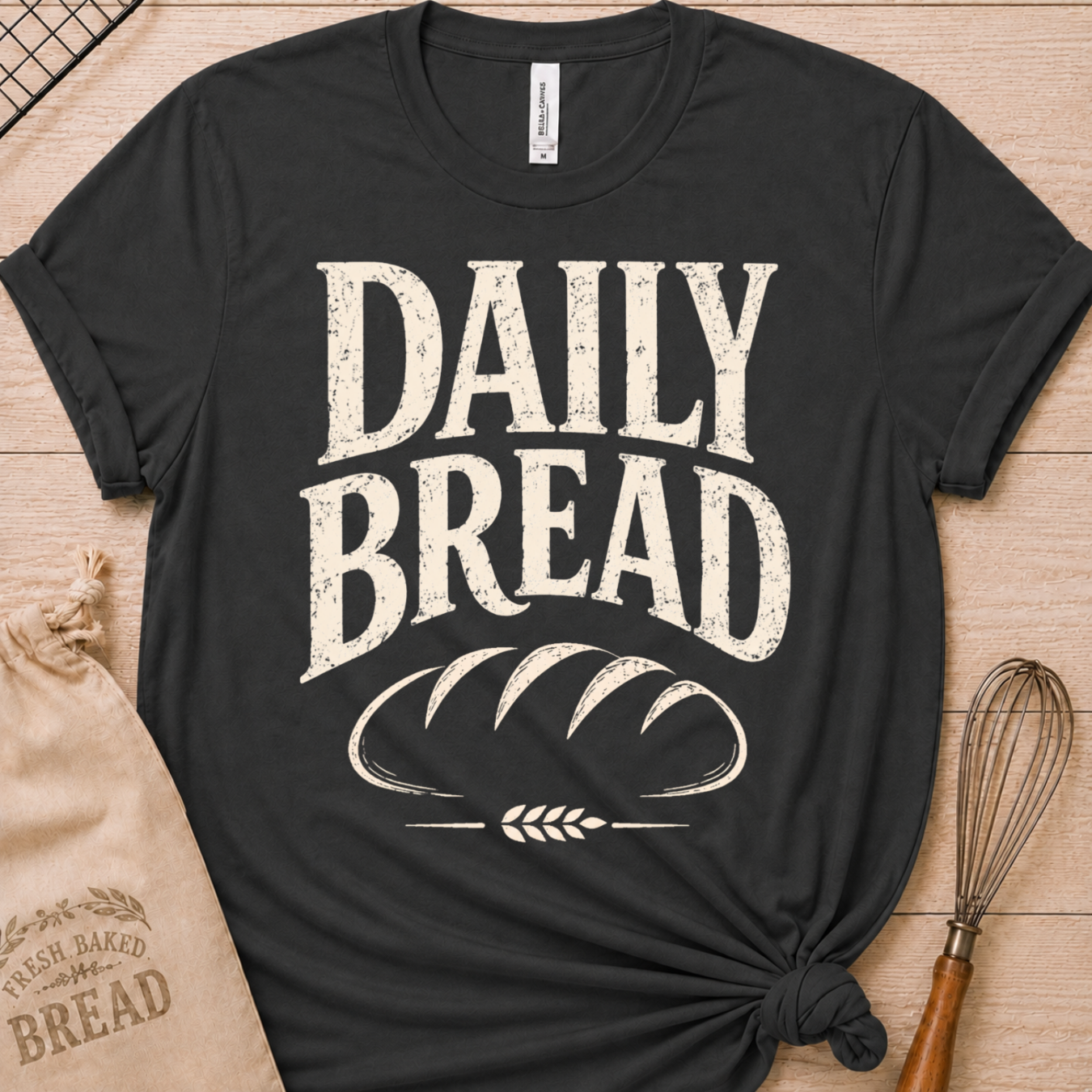 A dark heather grey short-sleeve graphic T-shirt laid flat on a light wood surface, printed with large distressed white text reading “DAILY BREAD” above a loaf-bread illustration. A kraft paper bag and whisk are styled props around the shirt.