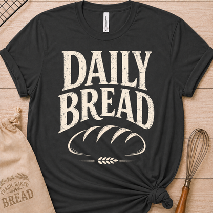 A dark heather grey short-sleeve graphic T-shirt laid flat on a light wood surface, printed with large distressed white text reading “DAILY BREAD” above a loaf-bread illustration. A kraft paper bag and whisk are styled props around the shirt.