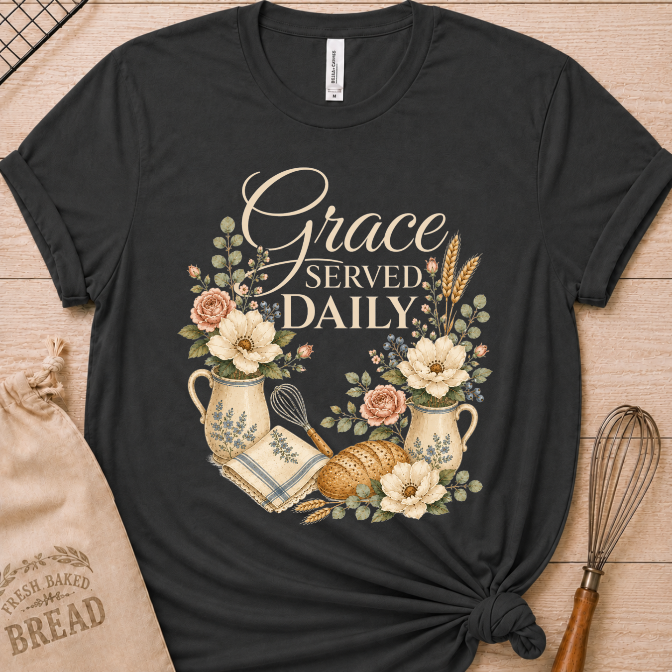 A black graphic t-shirt laid flat on a light wood background, featuring a vintage-style print that reads 'Grace SERVED DAILY' with flowers, bread, wheat, and kitchen utensils. A beige drawstring bag and a whisk are styled around the shirt.