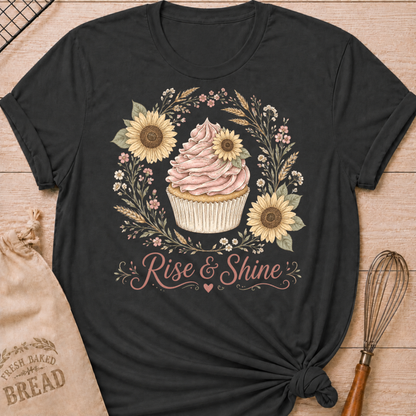 A black short-sleeve graphic T-shirt laid flat on a light wood surface, featuring a cupcake illustration surrounded by sunflowers and the words 'Rise & Shine.' A beige drawstring bread bag and a whisk are placed beside it as styling props.
