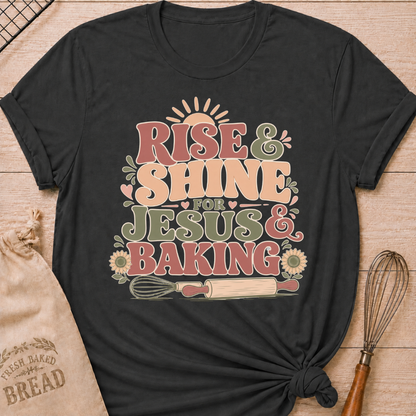 A dark heather grey graphic t-shirt styled with a knotted hem, laid flat on a light wood background with a drawstring bread bag and a whisk. The shirt features a bold retro groovy graphic that reads "RISE & SHINE FOR JESUS & BAKING" in pink, cream, and sage lettering, with a rising sun at the top, heart accents, sunflowers, and illustrations of a whisk and rolling pin at the bottom.
