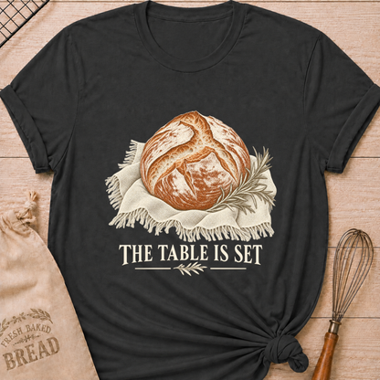 Dark Heather Grey women's relaxed fit tee with a rustic artisan loaf resting on softly draped linen with a rosemary sprig and The Table Is Set lettering