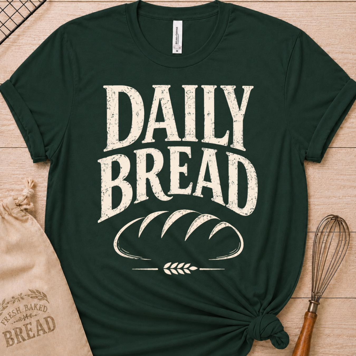 A dark green short-sleeve t-shirt with a distressed white graphic reading 'DAILY BREAD' and a loaf of bread illustration, styled flat-lay on a light wood surface with a burlap bag and whisk nearby.