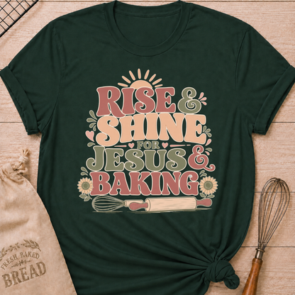 A forest green graphic t-shirt styled with a knotted hem, laid flat on a light wood background with a drawstring bread bag and a whisk. The shirt features a bold retro groovy graphic that reads "RISE & SHINE FOR JESUS & BAKING" in pink, cream, and sage lettering, with a rising sun at the top, heart accents, sunflowers, and illustrations of a whisk and rolling pin at the bottom.
