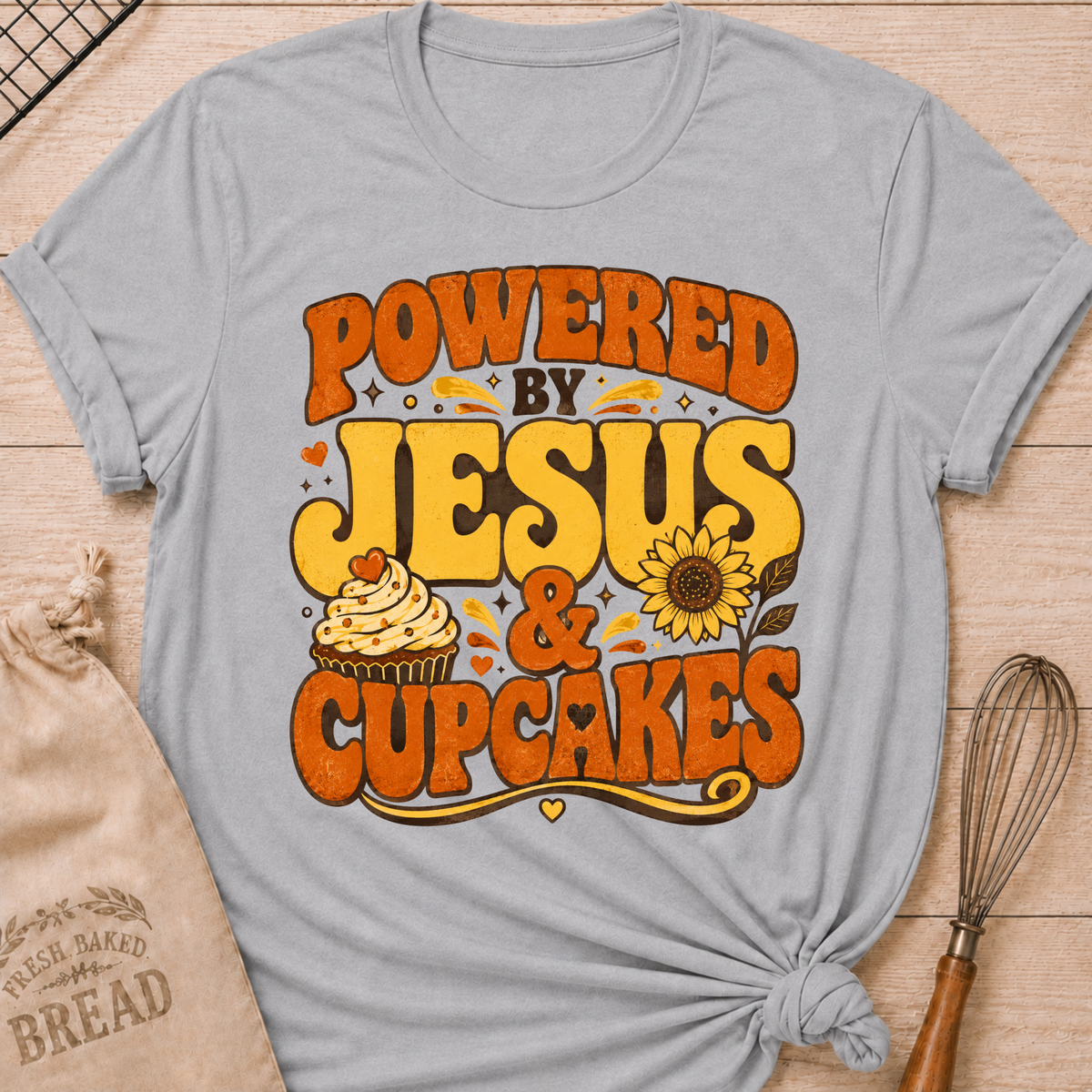 A light gray graphic t-shirt laid flat on a wooden surface, featuring a retro-style orange and yellow text design that reads 'POWERED BY JESUS & CUPCAKES' with a cupcake and sunflower illustration. A brown paper bread bag and a whisk are styled as props around the shirt.