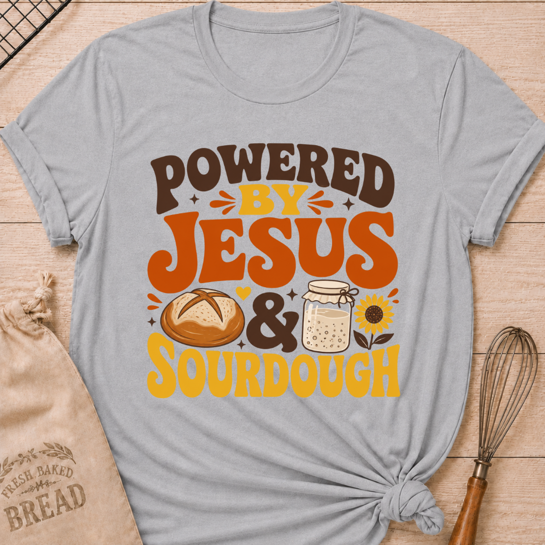 A Heather Grey graphic t-shirt laid flat on a light wood background, styled with a bread bag, whisk, and baking paper. The t-shirt features a graphic that reads 'POWERED BY JESUS & SOURDOUGH' in brown and orange lettering, accompanied by illustrations of a sourdough bread loaf, a mason jar of dough, and a sunflower.