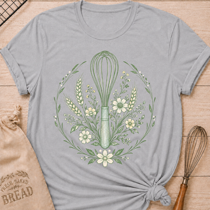 A heather grey graphic t-shirt styled with a knotted hem, laid flat on a light wood background with a drawstring bread bag and a whisk. The shirt features a detailed botanical illustration of a kitchen whisk centered within a circular wreath of wildflowers, wheat stalks, daisies, and delicate foliage, all rendered in sage green line art.