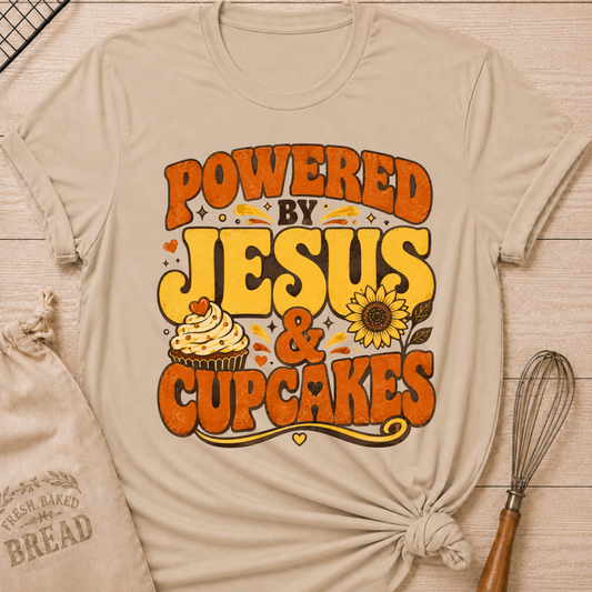 A beige graphic t-shirt with rolled sleeves, styled in a flat-lay on a light wood background with a drawstring bread bag, a whisk, and a small heart. The t-shirt features a retro-style graphic that reads 'POWERED BY JESUS & CUPCAKES' in orange and yellow lettering, accompanied by a cupcake and sunflower illustration.