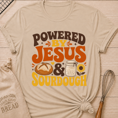 A beige graphic t-shirt with a 'POWERED BY JESUS & SOURDOUGH' design, styled in a flat-lay on a wooden surface with a bread bag, whisk, and baking pan. graphic t-shirt laid flat on a light wood background, styled with a bread bag, whisk, and baking paper. The t-shirt features a graphic that reads 'POWERED BY JESUS & SOURDOUGH' in brown and orange lettering, accompanied by illustrations of a sourdough bread loaf, a mason jar of dough, and a sunflower.