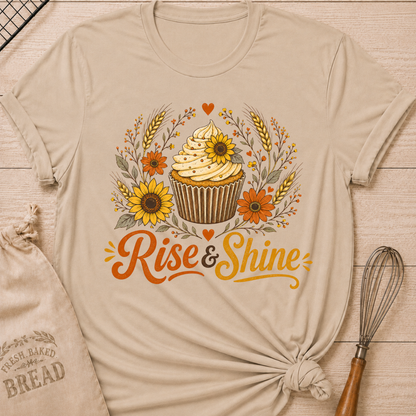 A heather stone graphic t-shirt styled with a knotted hem, laid flat on a light wood background with a drawstring bread bag and a whisk. The shirt features a warm, vintage-inspired illustration of a frosted cupcake surrounded by sunflowers, orange daisy-like flowers, wheat stalks, berry sprigs, and heart accents, with the words "Rise & Shine" written below in bold retro script in orange and golden yellow.