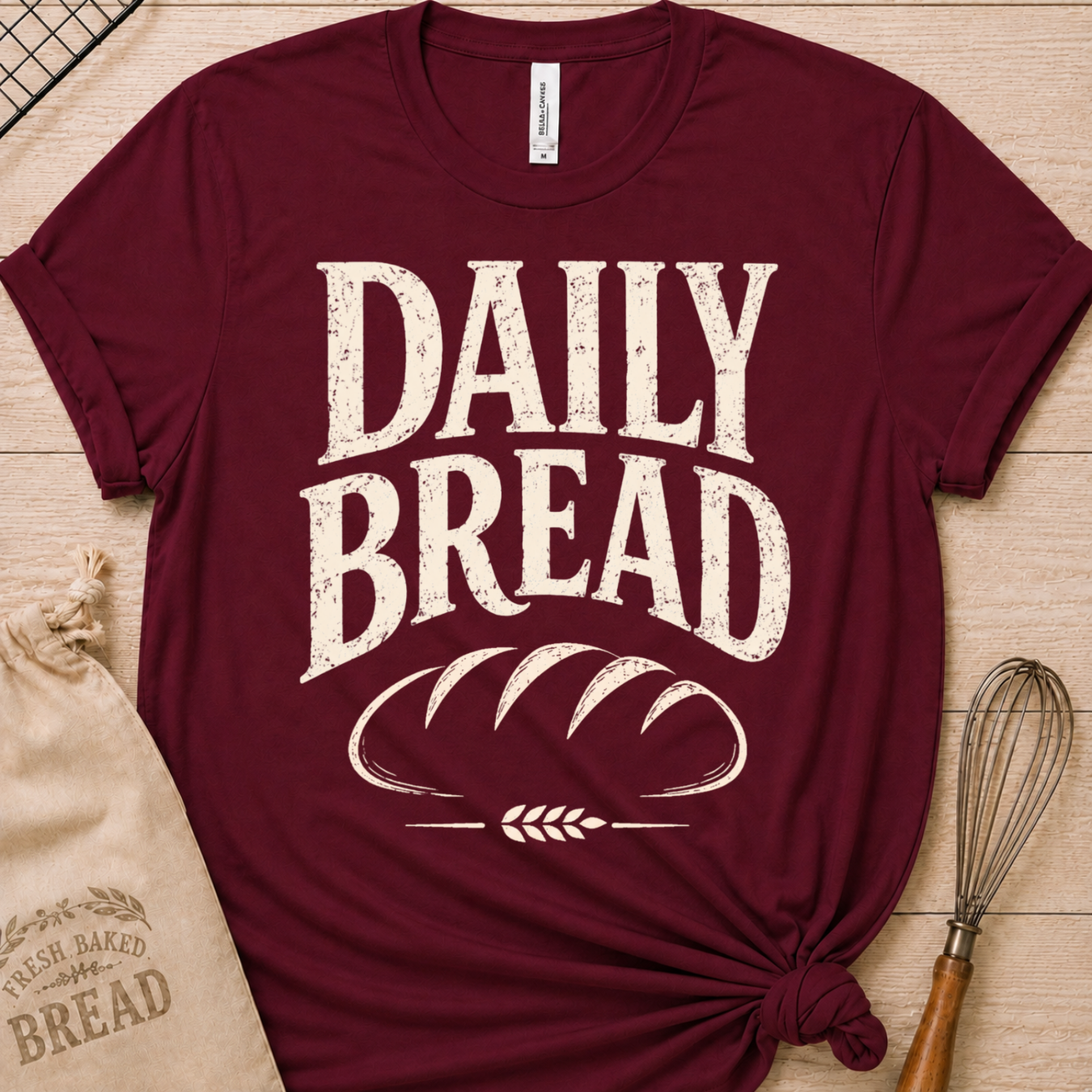 A maroon short-sleeve graphic T-shirt laid flat on a light wood surface, printed with large distressed white text reading “DAILY BREAD” above a loaf of bread illustration. A beige drawstring bag and whisk are placed around it as styling props.
