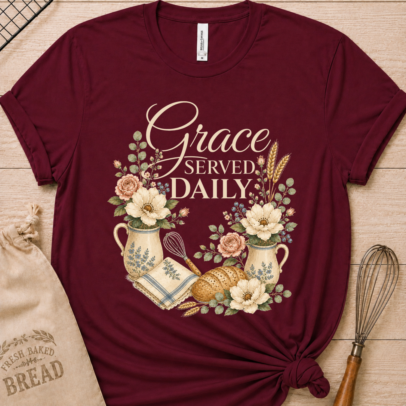 A maroon short-sleeve crewneck t-shirt with a 'Grace Served Daily' graphic, styled in a flat-lay on a light wood background with a burlap bag, whisk, and bread props.