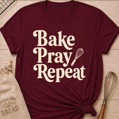 Maroon women's relaxed fit t-shirt with the words Bake, Pray, Repeat in playful vintage lettering and a classic kitchen whisk.
