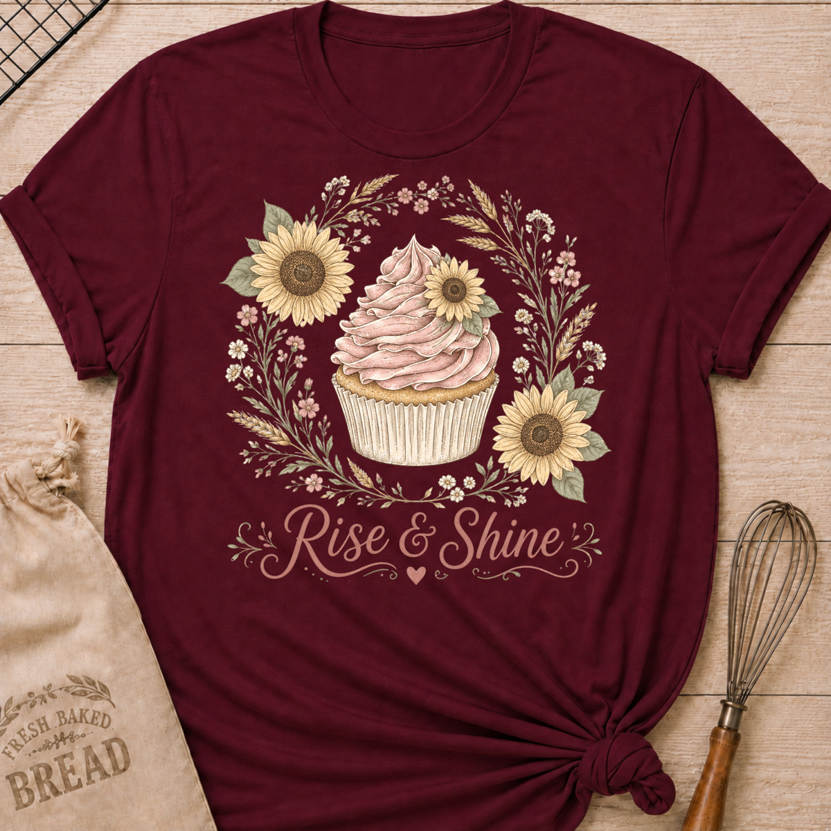 A maroon short-sleeve t-shirt with a cupcake and sunflower graphic, styled flat on a wooden surface with a burlap bread bag, wooden spoon, and wire whisk.