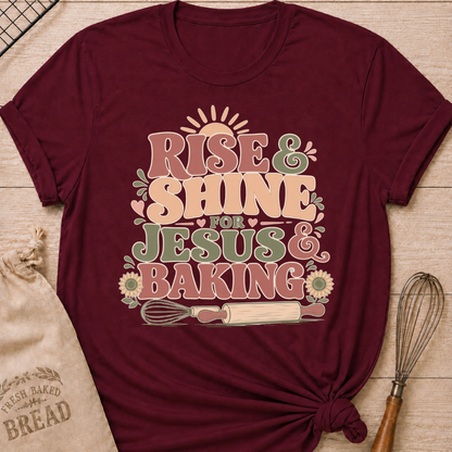 A maroon graphic t-shirt styled with a knotted hem, laid flat on a light wood background with a drawstring bread bag and a whisk. The shirt features a bold retro groovy graphic that reads "RISE & SHINE FOR JESUS & BAKING" in pink, cream, and sage lettering, with a rising sun at the top, heart accents, sunflowers, and illustrations of a whisk and rolling pin at the bottom.