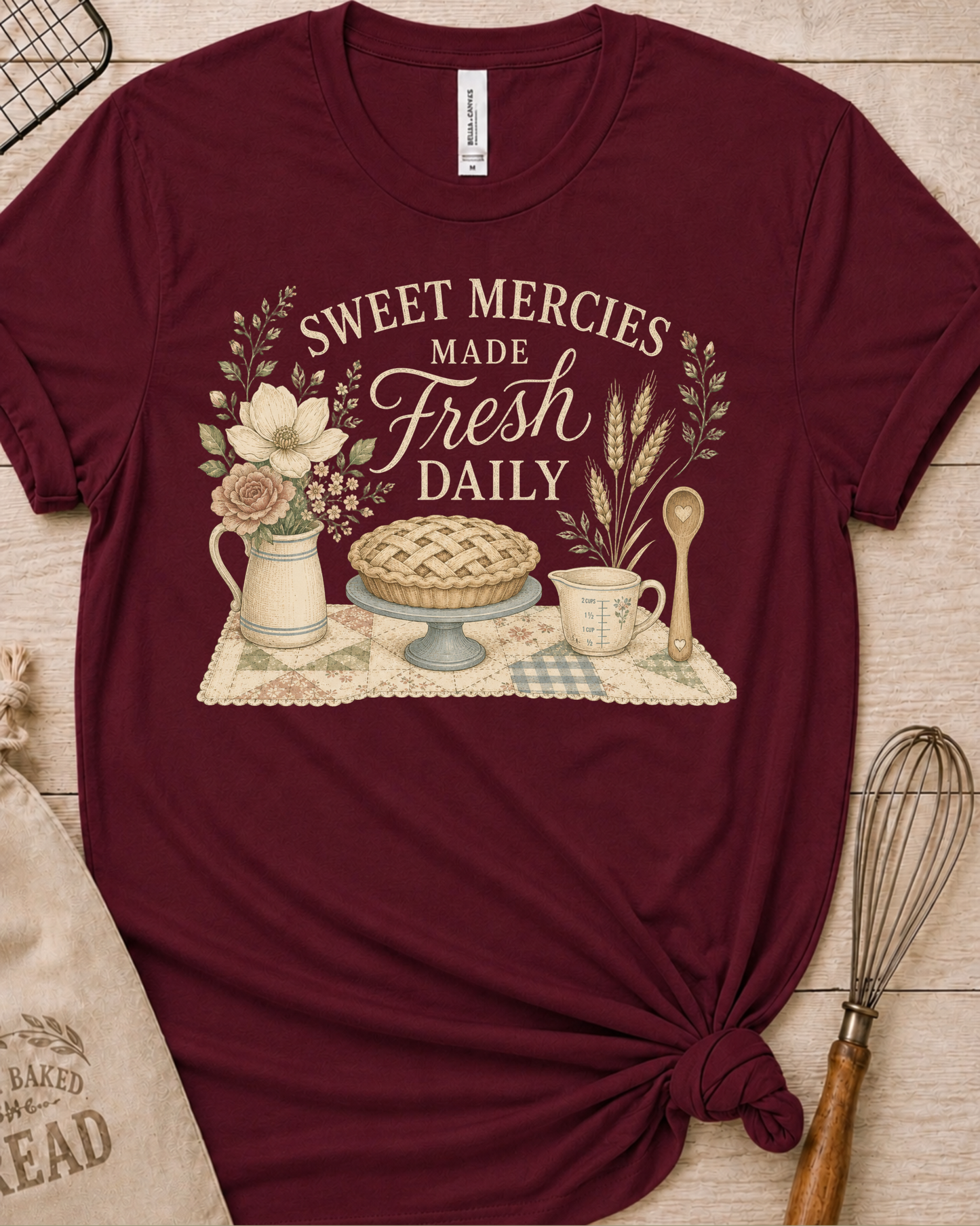 Maroon women's relaxed fit t-shirt reading Sweet Mercies Made Fresh Daily above a lattice-top pie on a cake stand with a flower pitcher, measuring cup, and wooden spoon.