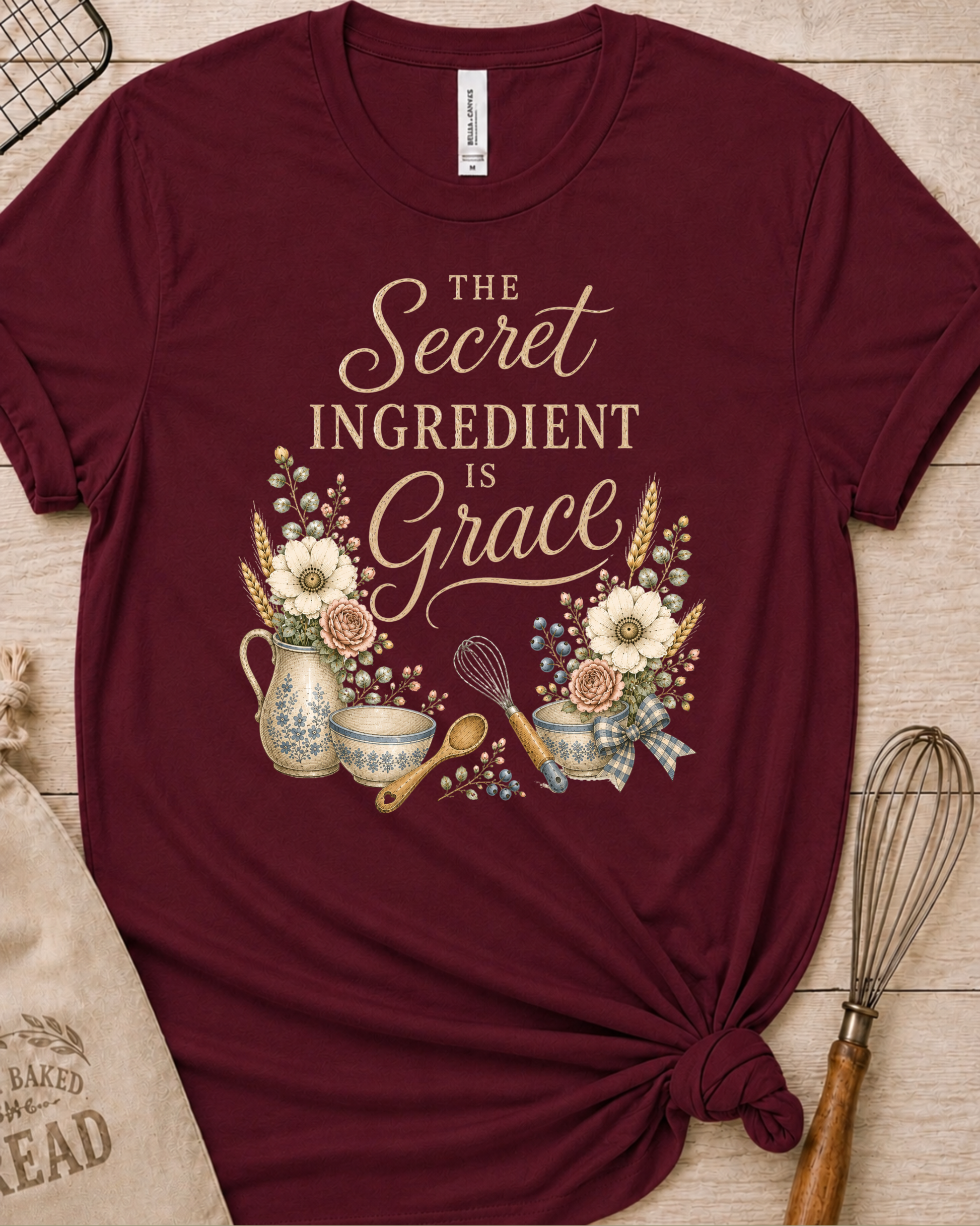 Maroon women's relaxed fit t-shirt reading The Secret Ingredient is Grace, framed by a floral pitcher, mixing bowls, a whisk, and a wooden spoon.
