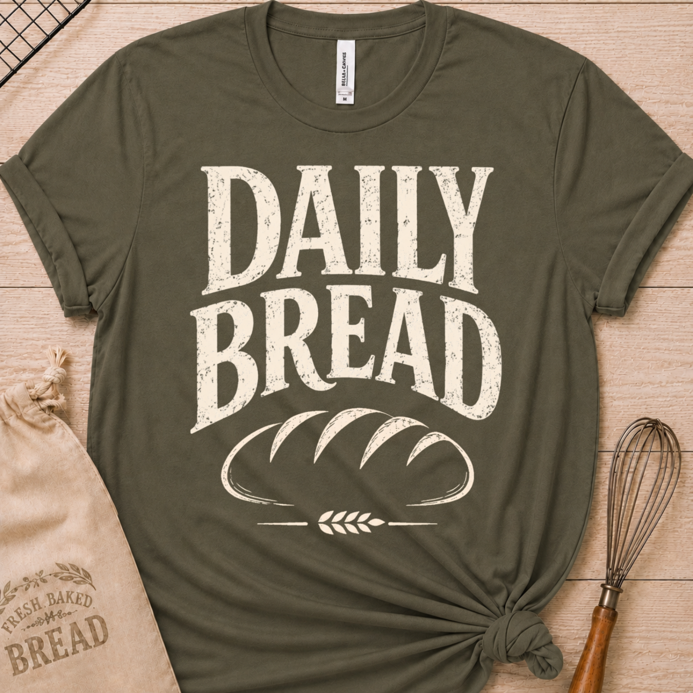 An olive green short-sleeve t-shirt with a distressed white graphic reading 'DAILY BREAD' and a loaf of bread design, styled flat-lay on a light wood background with a bread bag and whisk.