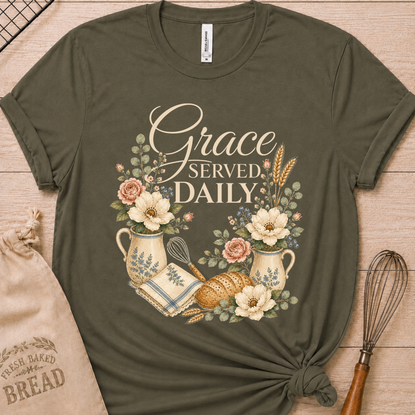 An olive green short-sleeve t-shirt with a 'Grace Served Daily' graphic, styled flat-lay on a light wood background with a burlap bread sack and a wire whisk.