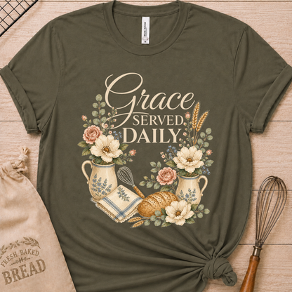 An olive green short-sleeve t-shirt with a 'Grace Served Daily' graphic, styled flat-lay on a light wood background with a burlap bread sack and a wire whisk.