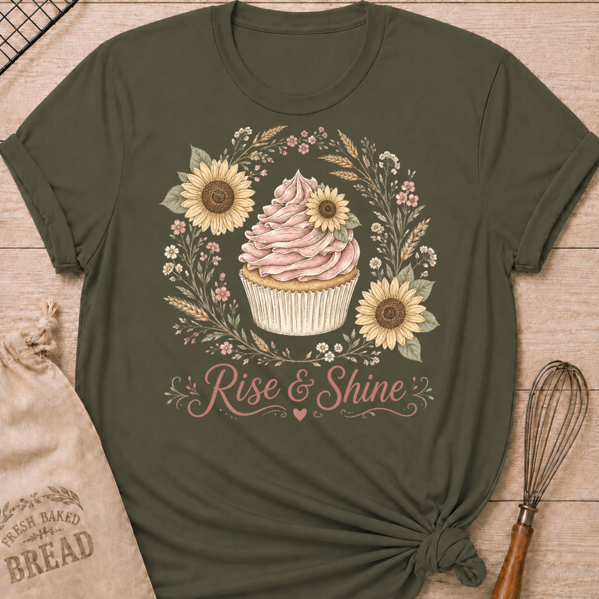 An olive green short-sleeve t-shirt with a cupcake graphic and the words 'Rise & Shine' printed on the front, styled flat on a light wood background with a bread bag and whisk nearby.