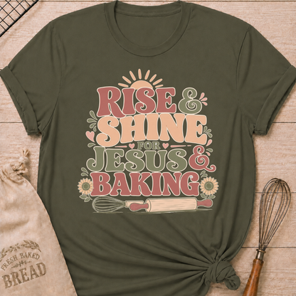 A military green graphic t-shirt styled with a knotted hem, laid flat on a light wood background with a drawstring bread bag and a whisk. The shirt features a bold retro groovy graphic that reads "RISE & SHINE FOR JESUS & BAKING" in pink, cream, and sage lettering, with a rising sun at the top, heart accents, sunflowers, and illustrations of a whisk and rolling pin at the bottom.