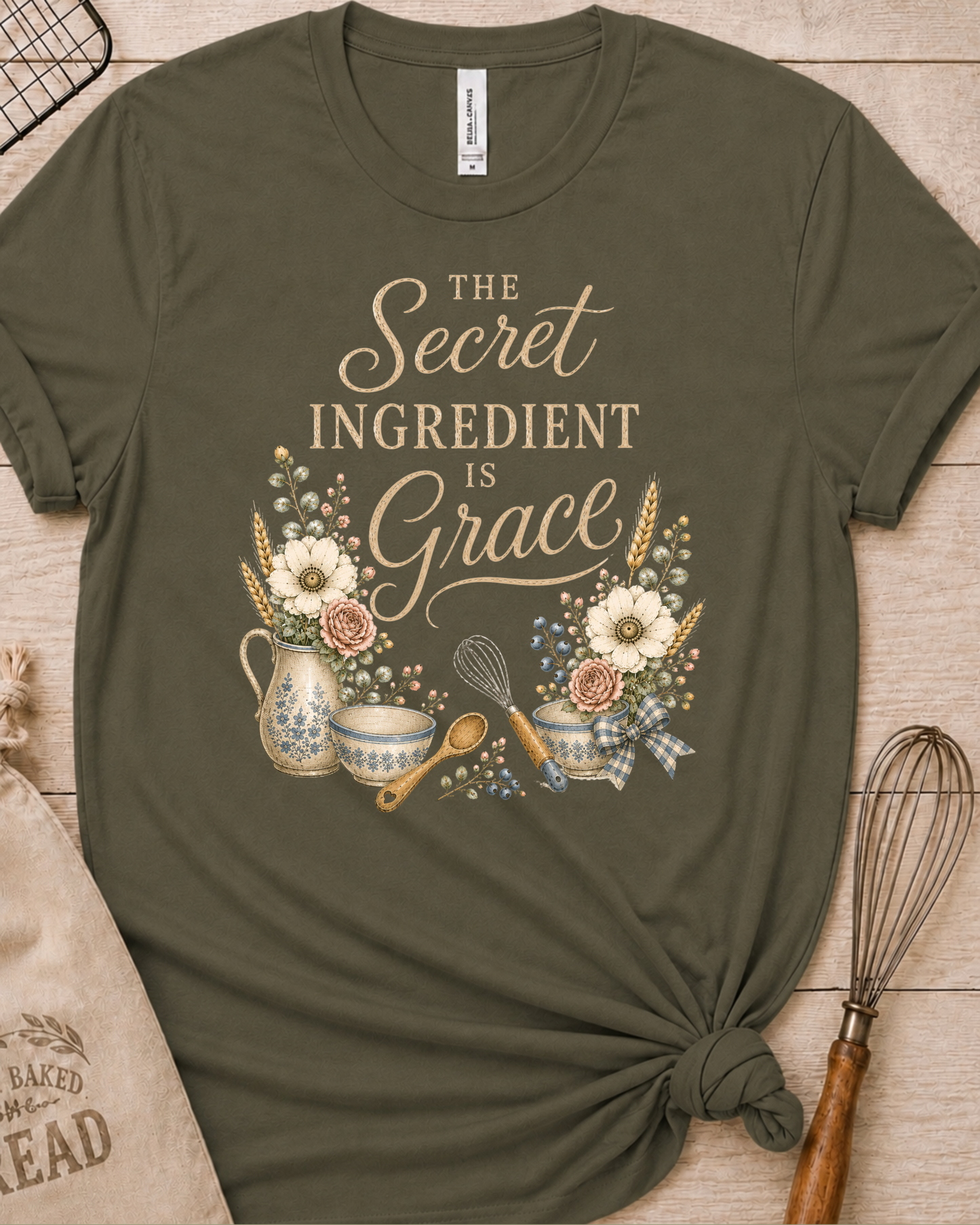 Military green women's relaxed fit t-shirt reading The Secret Ingredient is Grace, framed by a floral pitcher, mixing bowls, a whisk, and a wooden spoon.
