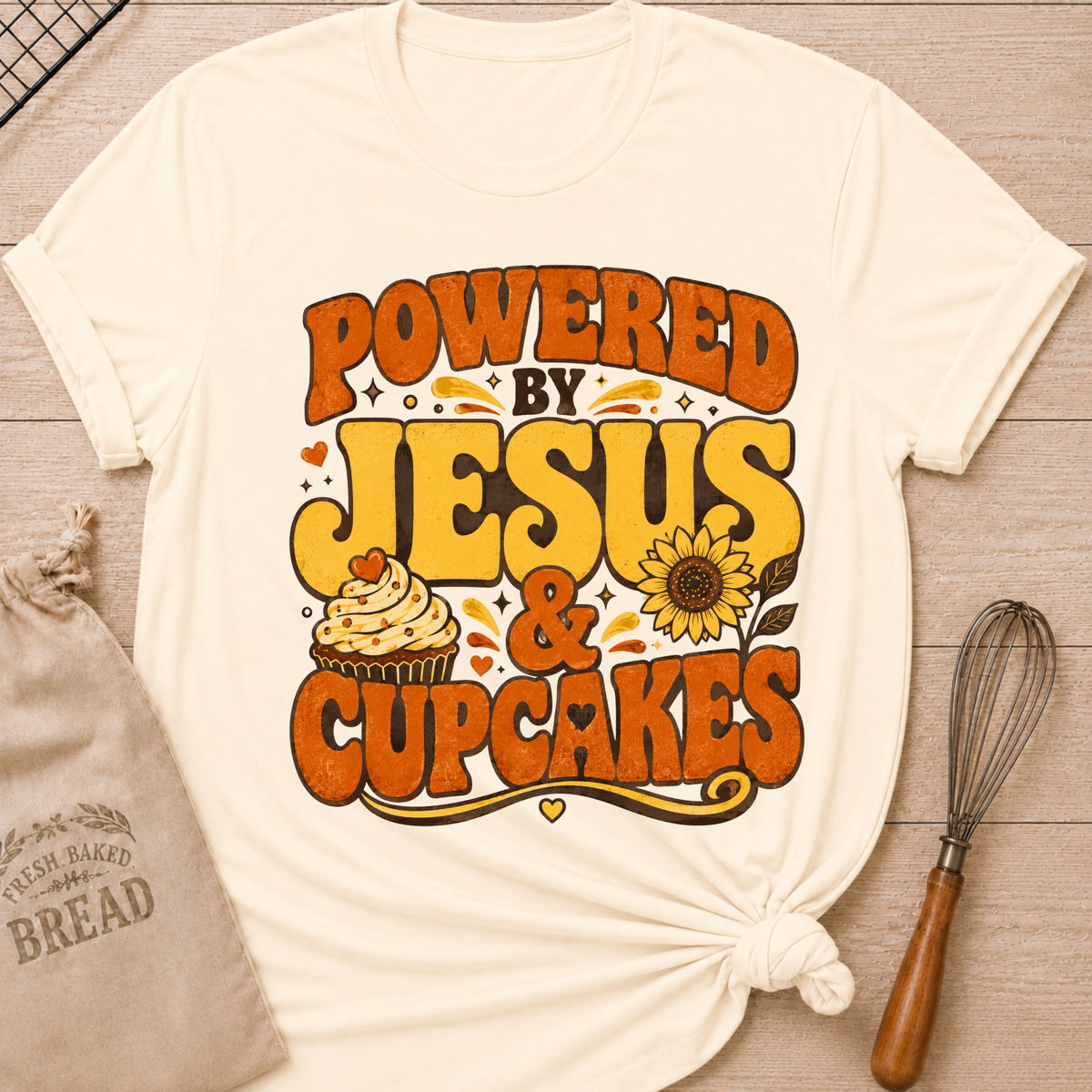 A cream-colored graphic t-shirt with a 'POWERED BY JESUS & CUPCAKES' design in orange and yellow, styled on a light wood background with a drawstring bag and a whisk. graphic t-shirt with rolled sleeves, styled in a flat-lay on a light wood background with a drawstring bread bag, a whisk, and a small heart. The t-shirt features a retro-style graphic that reads 'POWERED BY JESUS & CUPCAKES' in orange and yellow lettering, accompanied by a cupcake and sunflower illustration.