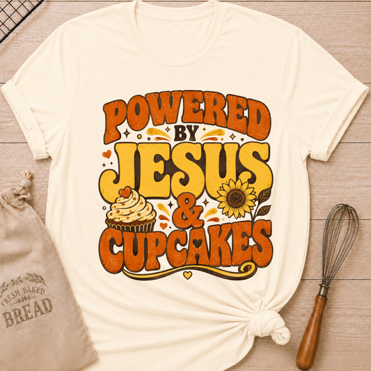 A cream-colored graphic t-shirt with a 'POWERED BY JESUS & CUPCAKES' design in orange and yellow, styled on a light wood background with a drawstring bag and a whisk. graphic t-shirt with rolled sleeves, styled in a flat-lay on a light wood background with a drawstring bread bag, a whisk, and a small heart. The t-shirt features a retro-style graphic that reads 'POWERED BY JESUS & CUPCAKES' in orange and yellow lettering, accompanied by a cupcake and sunflower illustration.