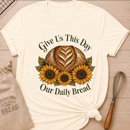 A cream-colored short-sleeve t-shirt with a graphic print of a loaf of bread, three sunflowers, and the text 'Give Us This Day Our Daily Bread', styled on a wooden background with a drawstring bag and a whisk. Women's relaxed fit natural ivory t-shirt laid flat on a light wood surface, featuring a faith-inspired graphic of a rustic sourdough boule with a heart-shaped wheat score surrounded by three golden sunflowers and deep olive green leaves, with the text "Give Us This Day Our Daily Bread" 