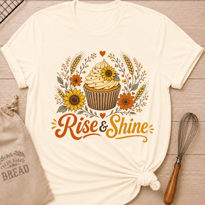 A natural, cream colored graphic t-shirt styled with a knotted hem, laid flat on a light wood background with a drawstring bread bag and a whisk. The shirt features a warm, vintage-inspired illustration of a frosted cupcake surrounded by sunflowers, orange daisy-like flowers, wheat stalks, berry sprigs, and heart accents, with the words "Rise & Shine" written below in bold retro script in orange and golden yellow.