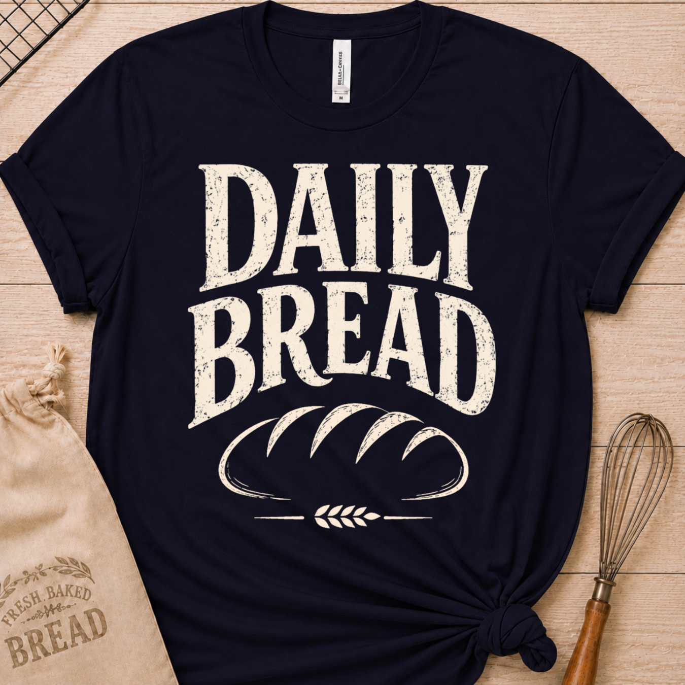A navy blue graphic t-shirt with a distressed white print reading 'DAILY BREAD' and a loaf of bread illustration, styled in a flat lay on a wooden surface with a burlap bag and whisk.