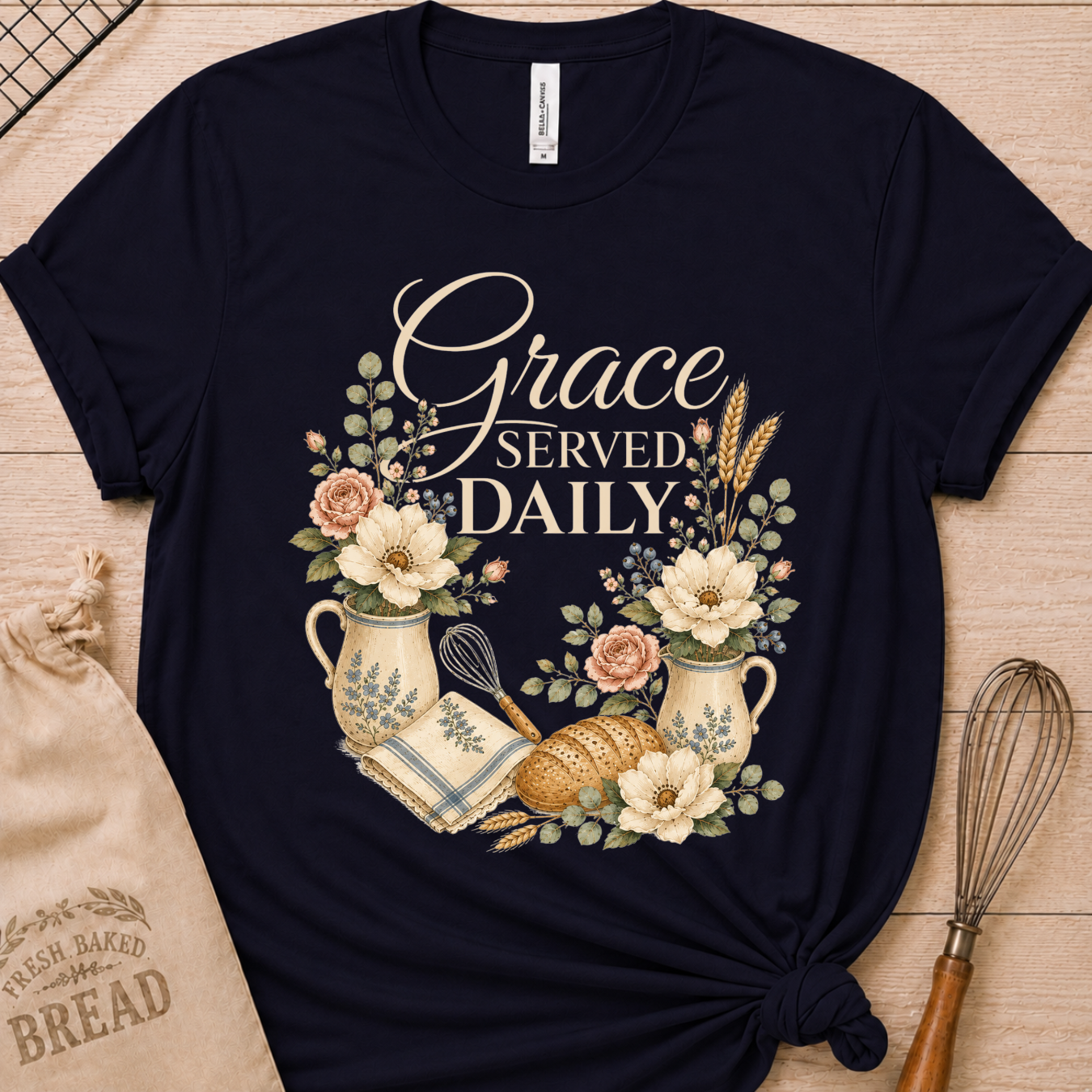 A navy blue graphic t-shirt laid flat on a light wood background, styled with a bread sack and a whisk. The t-shirt features a central graphic with the words 'Grace SERVED DAILY' in white script and serif fonts, surrounded by illustrations of white and pink flowers, green leaves, a cream pitcher, a whisk, and a loaf of bread.