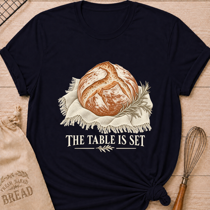 Navy women's relaxed fit tee with a rustic artisan loaf resting on softly draped linen with a rosemary sprig and The Table Is Set lettering