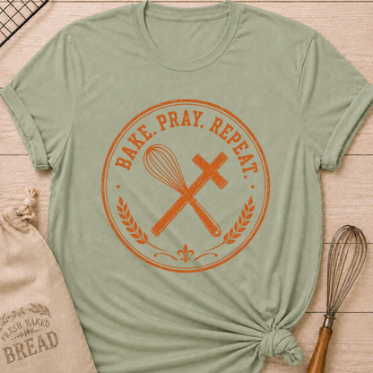 Sage green women's relaxed fit t-shirt with the words Bake. Pray. Repeat. inside a vintage bakery emblem with a classic whisk, a rolling pin, and wheat details.