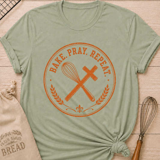 Sage green women's relaxed fit t-shirt with the words Bake. Pray. Repeat. inside a vintage bakery emblem with a classic whisk, a rolling pin, and wheat details.