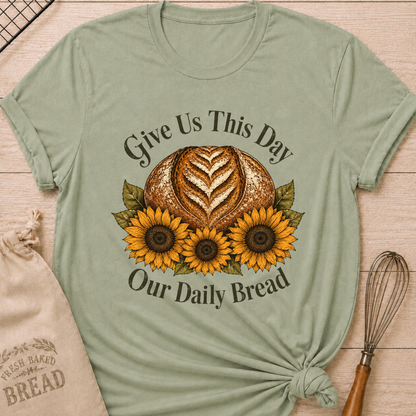 A sage green short-sleeve t-shirt with a rustic graphic of a loaf of bread and three sunflowers, with the text 'Give Us This Day Our Daily Bread'. The shirt is styled in a flat-lay on a light wood background with a beige drawstring bag and a whisk. Women's relaxed fit sage green t-shirt laid flat on a light wood surface, featuring a faith-inspired graphic of a rustic sourdough boule with a heart-shaped wheat score surrounded by three golden sunflowers and deep olive green leaves