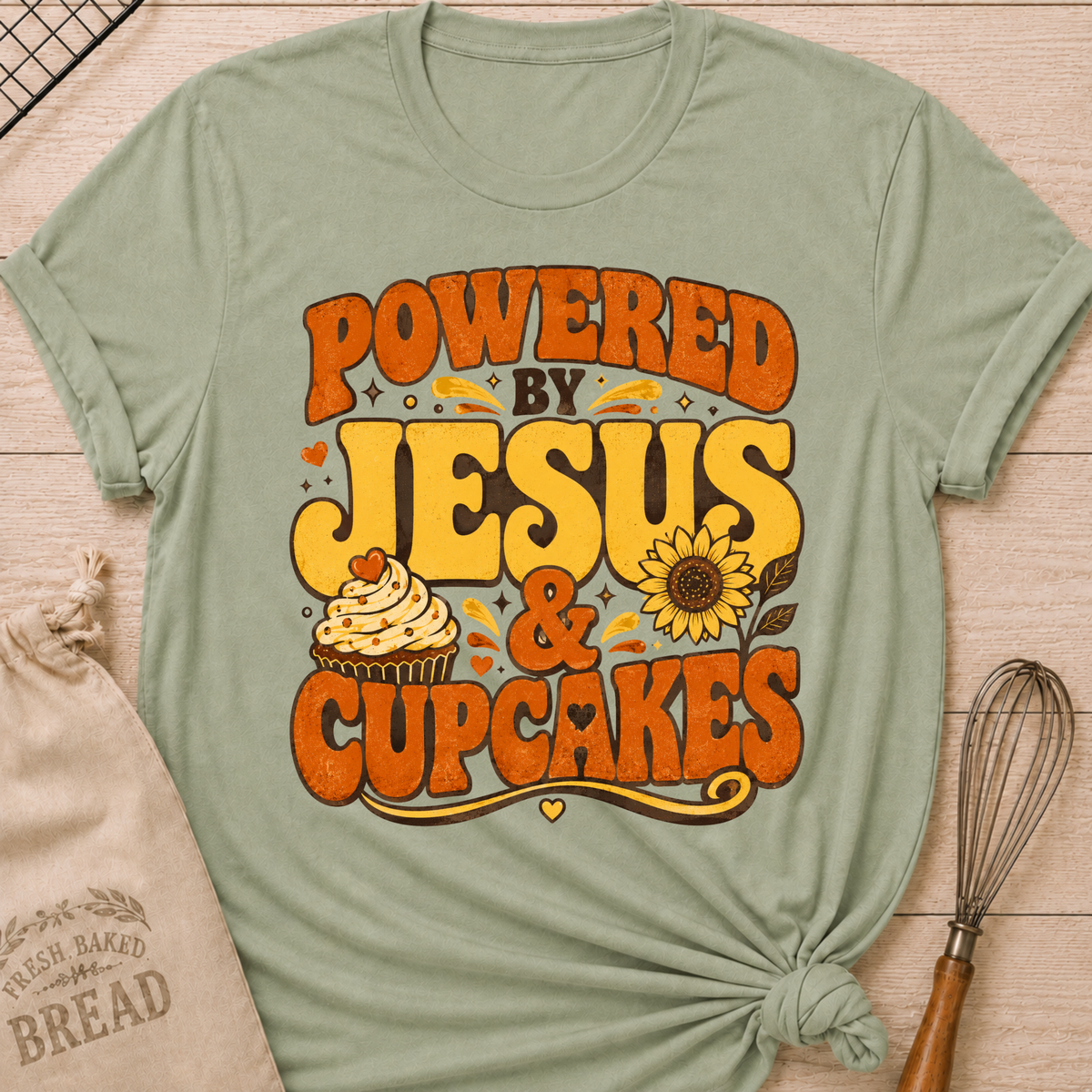A sage green short-sleeve t-shirt with a distressed retro graphic that reads 'POWERED BY JESUS & CUPCAKES' in orange and yellow lettering, featuring a cupcake and sunflower illustration. The shirt is styled in a flat-lay on a light wood background with a knotted hem, accompanied by a burlap sack and a whisk.