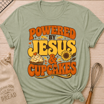 A sage green short-sleeve t-shirt with a distressed retro graphic that reads 'POWERED BY JESUS & CUPCAKES' in orange and yellow lettering, featuring a cupcake and sunflower illustration. The shirt is styled in a flat-lay on a light wood background with a knotted hem, accompanied by a burlap sack and a whisk.
