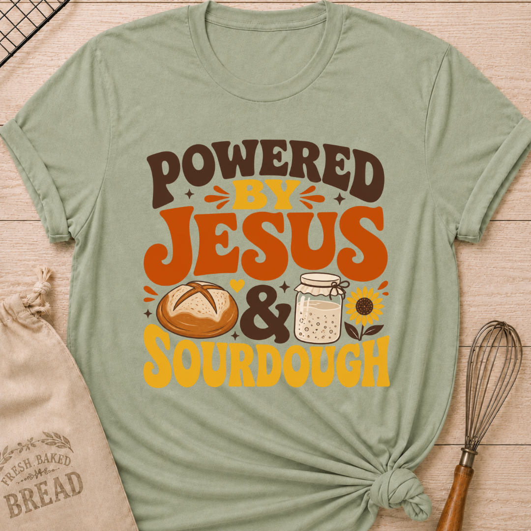 A sage green graphic t-shirt laid flat on a light wood background, styled with a bread bag, whisk, and baking paper. The t-shirt features a graphic that reads 'POWERED BY JESUS & SOURDOUGH' in brown and orange lettering, accompanied by illustrations of a sourdough bread loaf, a mason jar of dough, and a sunflower.